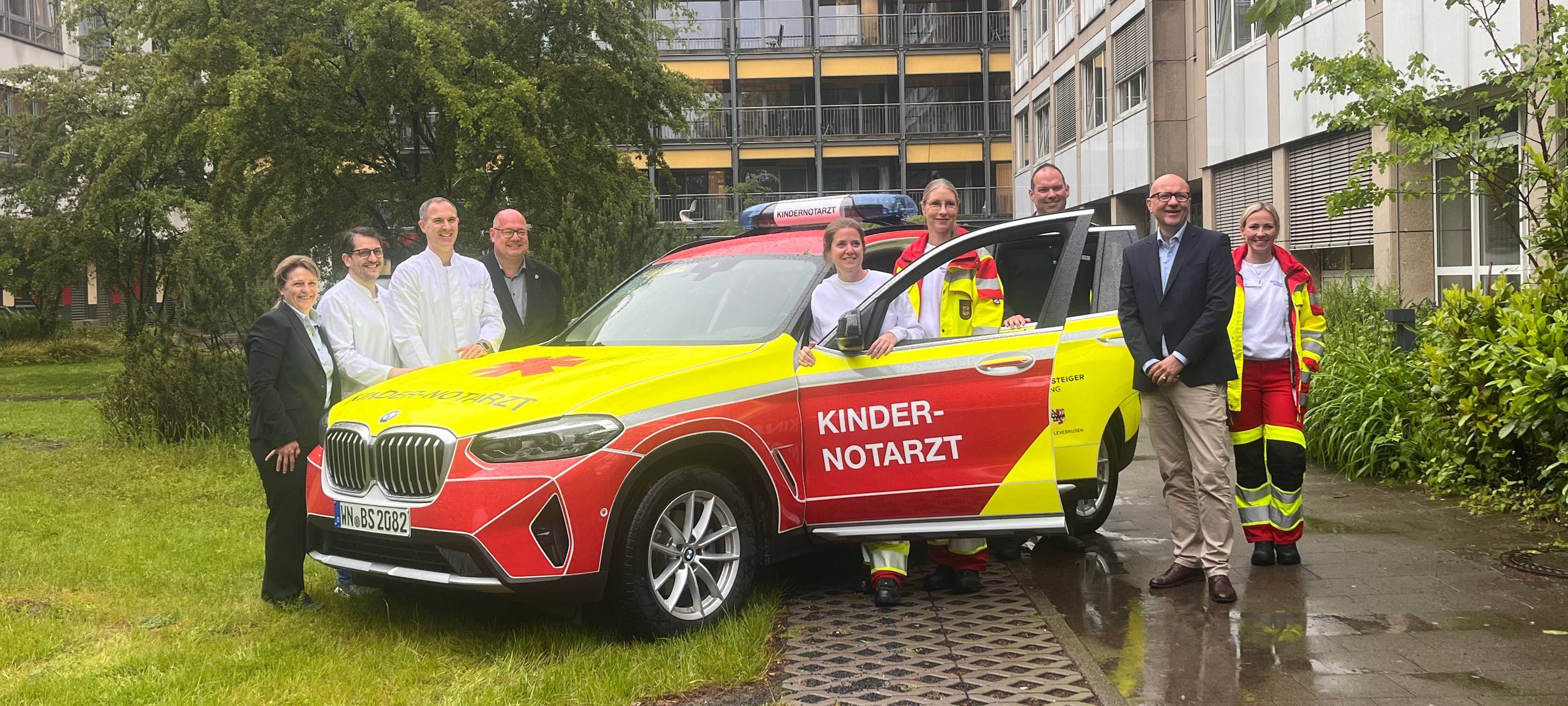 Das Kinder Notfall Team steht vor dem rot-gelben Sonderfahrzeug, was für die Einsätze unterwegs ist.