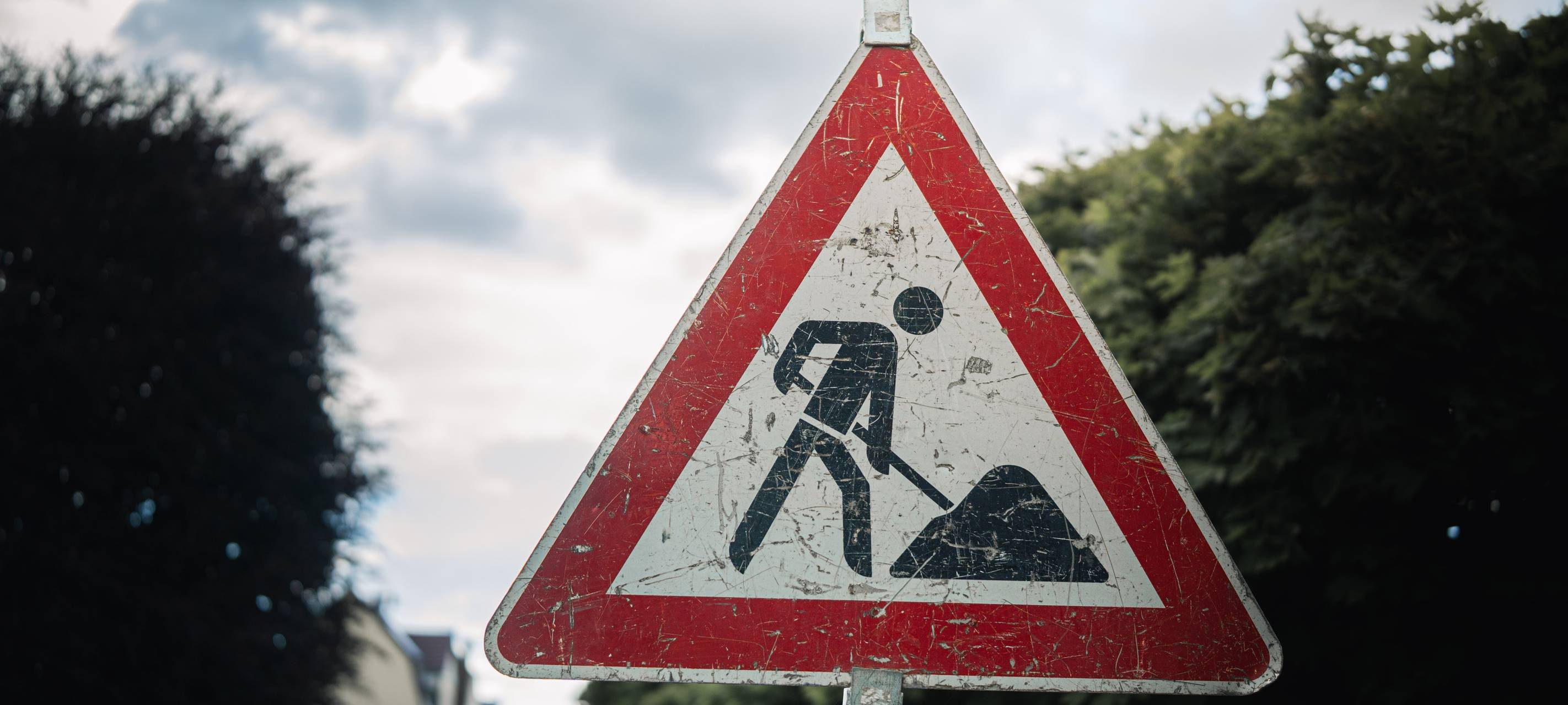 Dreieckiges Verkehrsschild „Baustelle“ mit Arbeiter-Symbol an einer Straße, im Hintergrund Bäume und unscharfe Häuser.