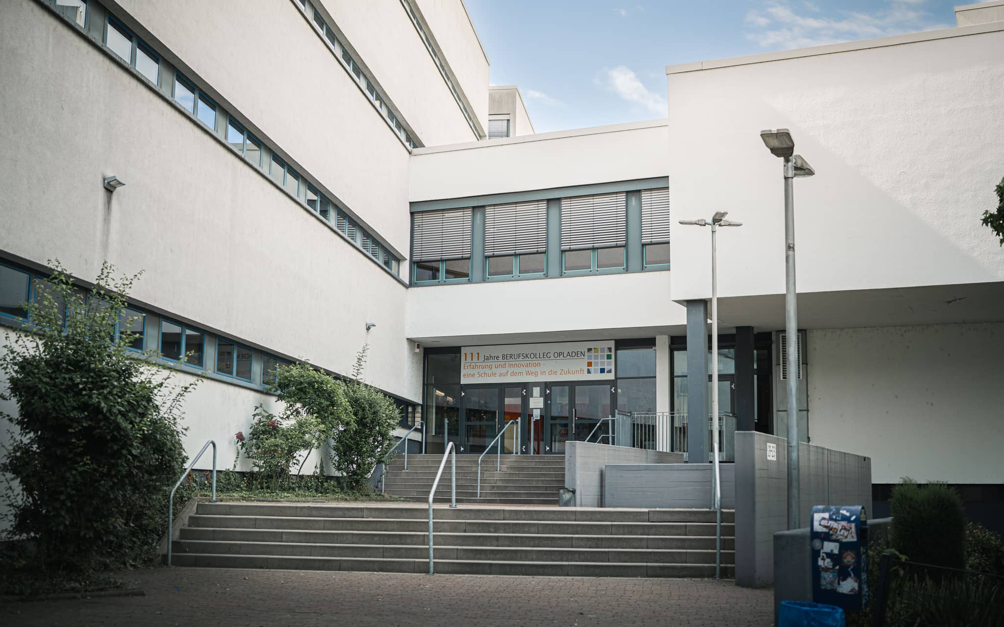 Eingangsbereich des Berufskollegs Opladen mit Treppenaufgang, Geländern und dem Schulgebäude im Hintergrund.